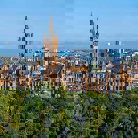 Panoramic view of the historic University of Glasgow building, with trees in the foreground and city skyline behind.