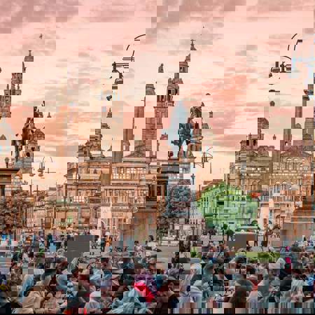 George Square in Glasgow at sunset, with a crowd of people gathered in the foreground and the illuminated City Chambers building and statues in the background.