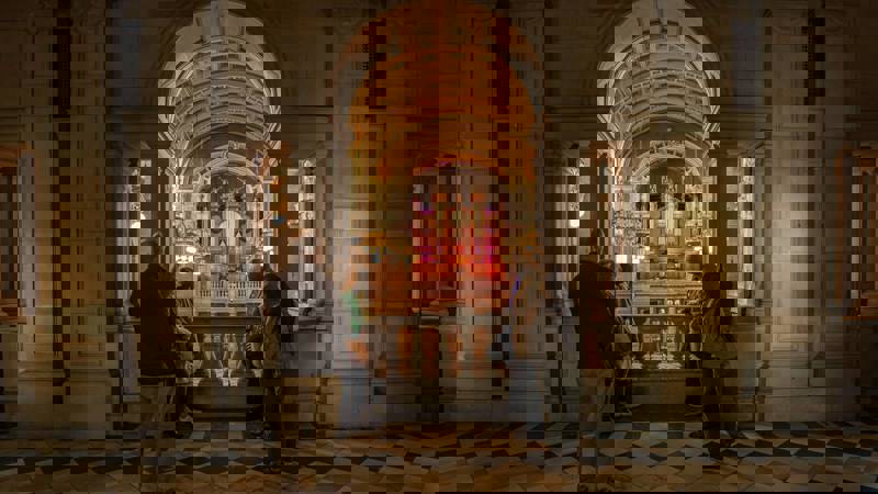 People stand on a balcony with a checkered floor, looking at a large, ornate organ in a grand hall.