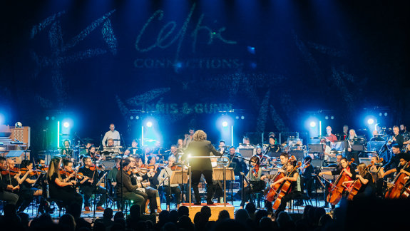 An orchestra performing on stage under blue spotlights, with a conductor facing the musicians and the Celtic Connections logo projected behind.