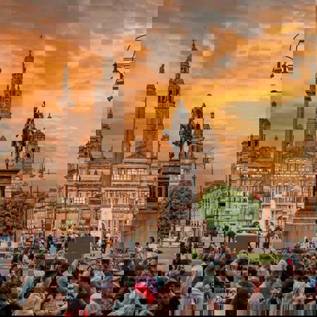 A vibrant photo of a crowd of people gathered in Glasgow's George Square at sunset. In the background, the Glasgow City Chambers stands under a beautiful orange and cloudy sky.
