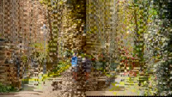 Two people walk along a tree-lined path next to an old stone building on a sunny day.