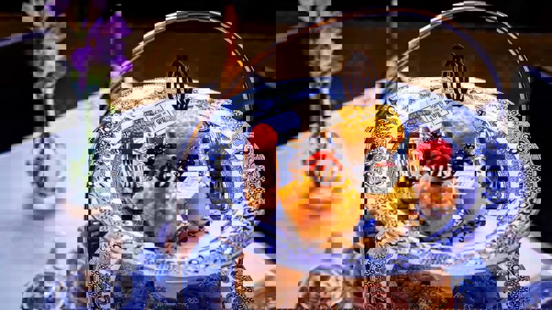 Close-up of delicate pastries, including muffins and chocolate-decorated eclairs, on a blue and white patterned plate with a "Mackintosh at The Willow" white chocolate tag.