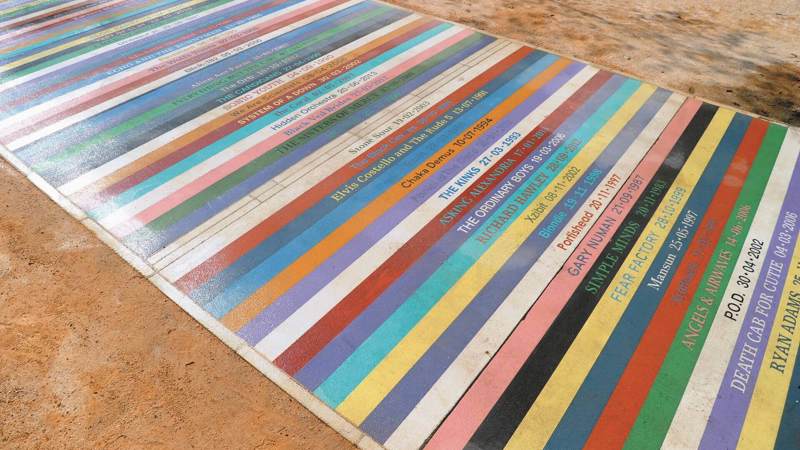 A ground-level artwork with colourful stripes, each labelled with a musician's name and a date.