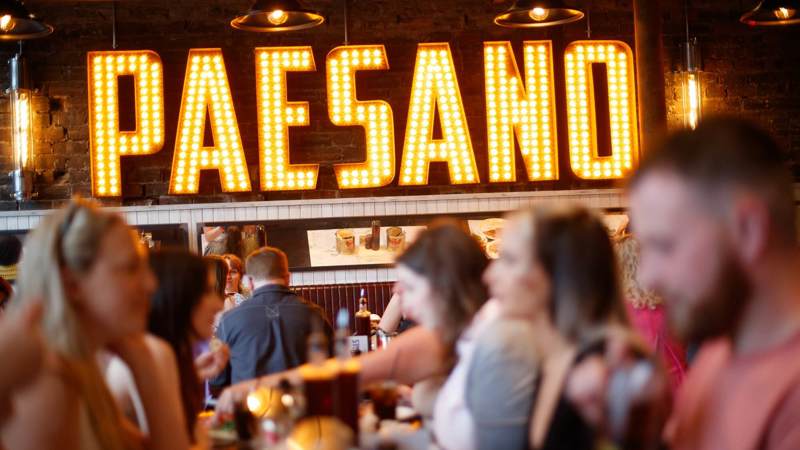 The interior of a busy Italian restaurant, with a large, lit-up 'PAESANO' sign on the wall behind people dining at tables.