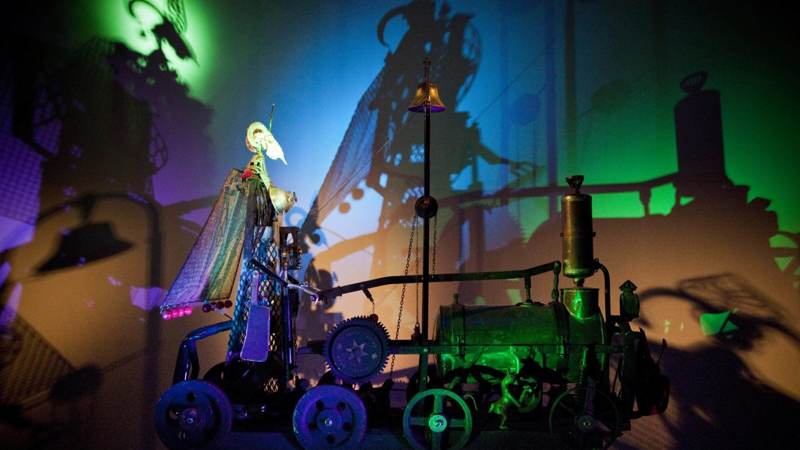 A kinetic sculpture from Sharmanka in Glasgow, with a strange figure on a wheeled machine casting dramatic shadows on a colourful wall.