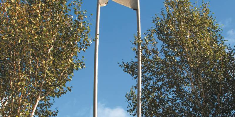 A tall, abstract metal sculpture resembling a safety pin stands between two birch trees against a bright blue sky with a bird perched on top.