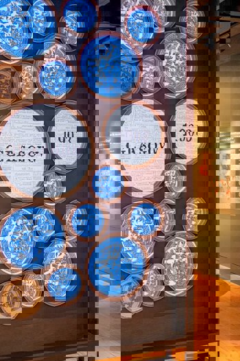 An interior shot of an exhibition with large circular art pieces on a wall, some with text like "CITY GLASGOW 1903".