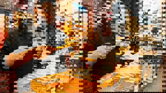 A person with a beard and glasses uses yellow tongs to place a pastry into a paper bag. They stand behind a counter displaying trays of cinnamon rolls and other baked goods inside Ottoman Coffee House.