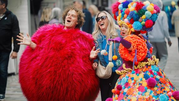 Two street performers in vibrant, colourful costumes, one in a red fuzzy suit and another in a multi-coloured pom-pom dress, pose with a woman.