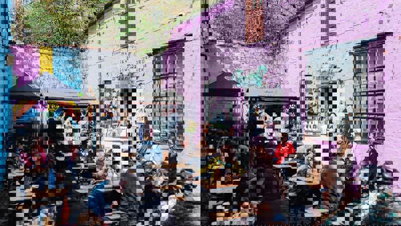 A bustling outdoor market scene with many people sitting at picnic tables in a courtyard with bright pink and yellow walls.