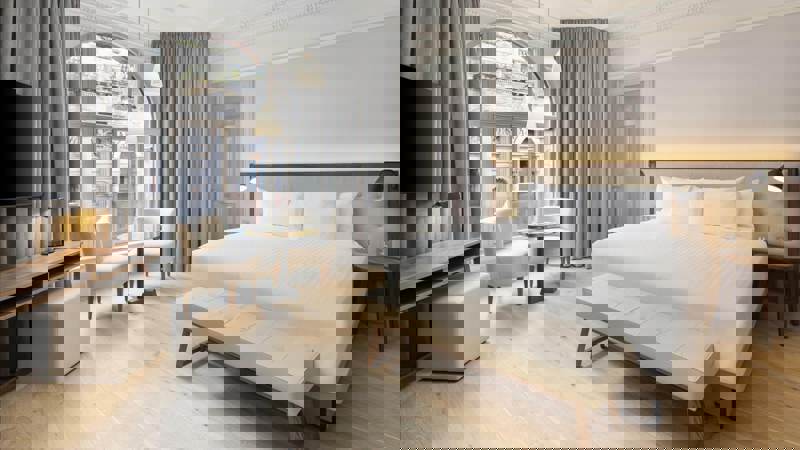 Stylish guest room at AC Hotel Glasgow with a large bed, modern lighting, light wooden floors, and a seating area beside tall arched windows overlooking the city.