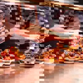 A chef's hands are plating dishes of roasted corn on the cob and other food on a metal counter in a restaurant kitchen.
