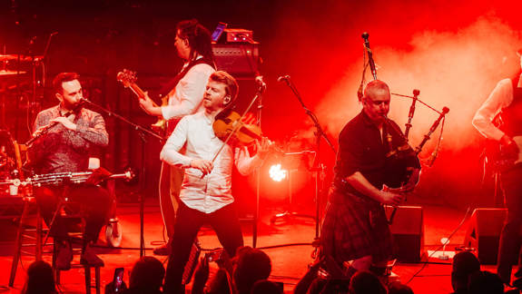 A band performs on a stage under red lighting. The musicians are playing traditional instruments, including a fiddle, a woodwind instrument, and bagpipes.