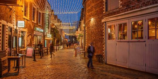 A cobblestone lane in Glasgow, Ashton Lane, lit by hanging fairy lights and lanterns, with people walking along it at dusk.