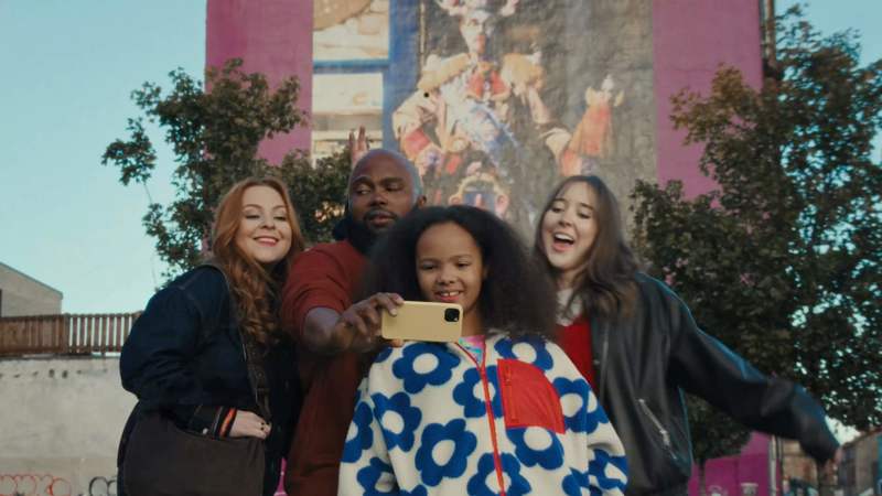 A group of four smiling friends take a selfie together outdoors. They stand before a pink building featuring a large mural, surrounded by green trees under a clear sky.