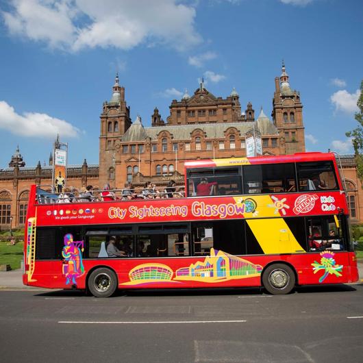 A bright red double-decker city sightseeing bus in Glasgow, with passengers on the top deck, passing the grand Kelvingrove Art Gallery and Museum.
