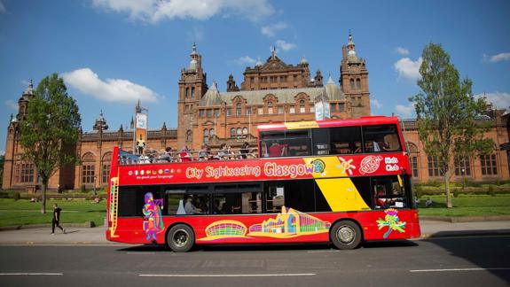 A bright red double-decker city sightseeing bus in Glasgow, with passengers on the top deck, passing the grand Kelvingrove Art Gallery and Museum.