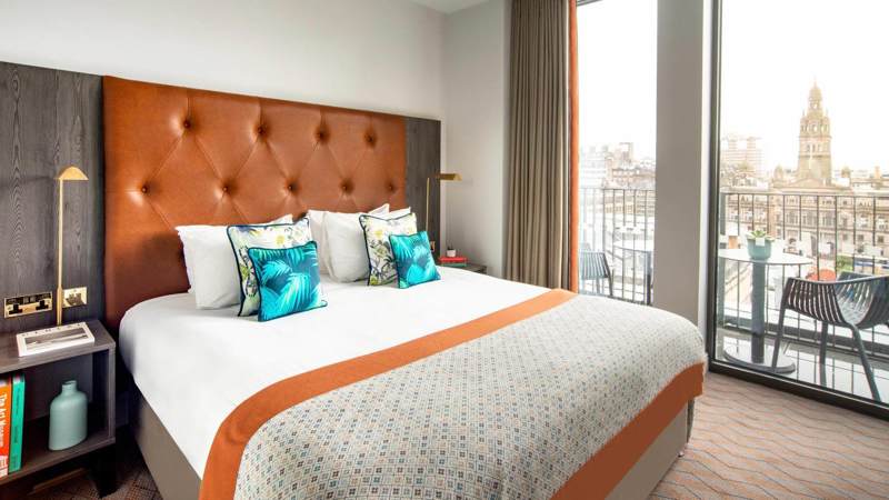 Modern apartment-style room at Native Glasgow with a king-size bed, large windows overlooking George Square, and stylish mid-century furnishings in warm tones.