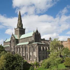 The grand exterior of Glasgow Cathedral with its spire reaching into a blue sky with white clouds, surrounded by green trees.
