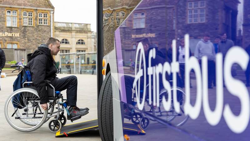 A person in a wheelchair is using the ramp to board a bus with "First Bus" written in large white letters on its purple side.