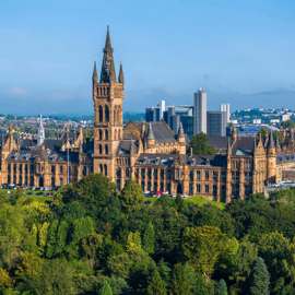Panoramic view of the historic University of Glasgow building, with trees in the foreground and city skyline behind.