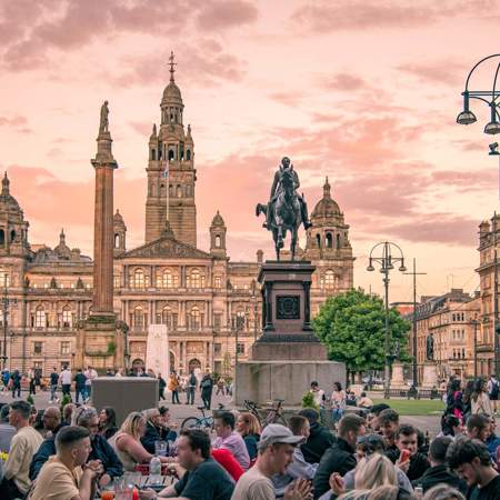 George Square in Glasgow at sunset, with a crowd of people gathered in the foreground and the illuminated City Chambers building and statues in the background.