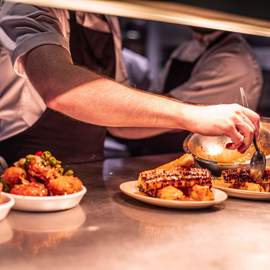 A chef's hands are plating dishes of roasted corn on the cob and other food on a metal counter in a restaurant kitchen.